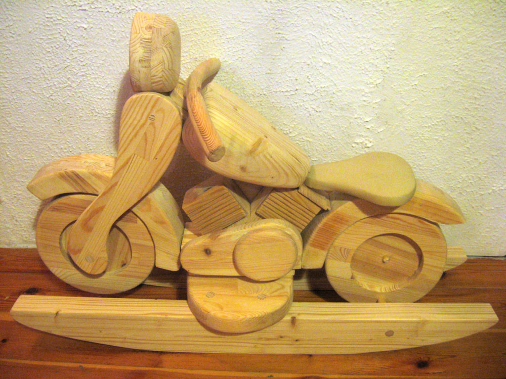 Drewniany motocykl Harley na biegunach, wykonany z jasnego drewna sosnowego, widoczny na tle białej, teksturowanej ściany i drewnianej podłogi.